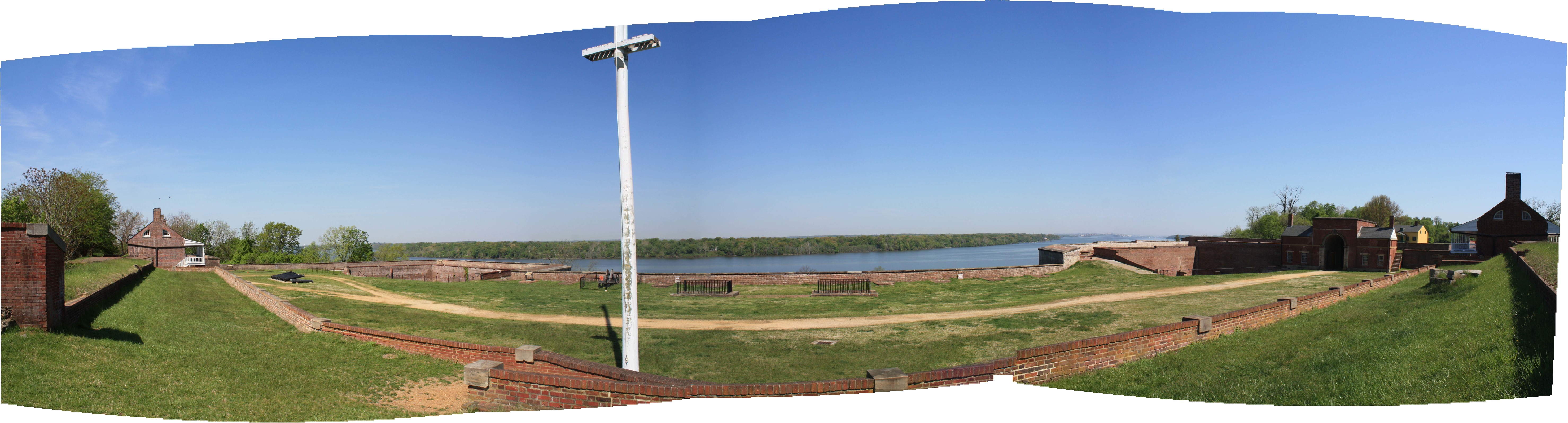 Wide panoramic view towards a river, with the brick walls, earthworks, and structures of a coastal fort in the foreground. 