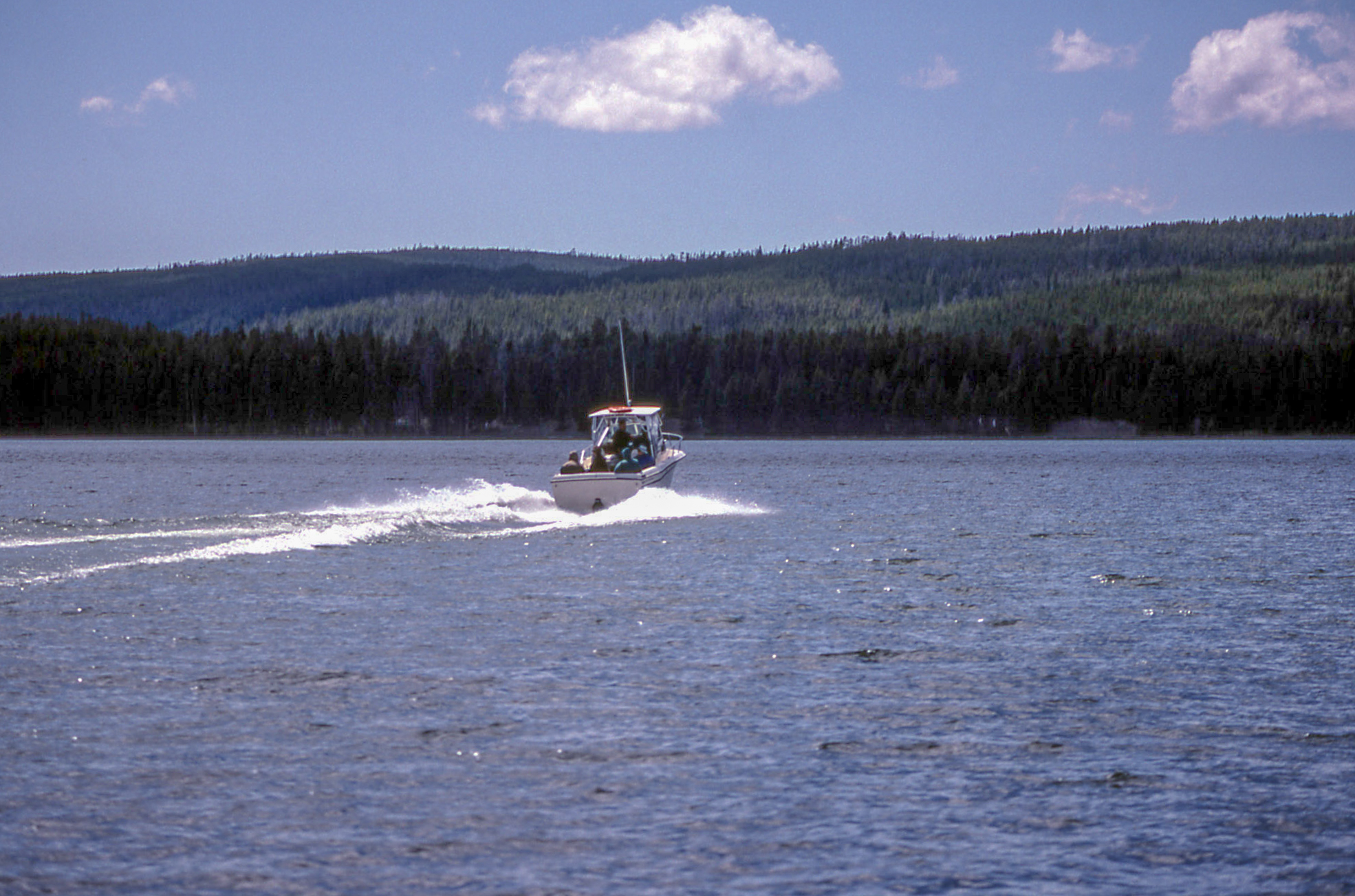 A fishing boat is powering up on a lake going away from the camera with low forested hills in the foreground.