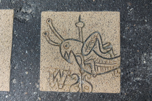 This is a close up of a part of the mosaic for the first public school, the Latin School. It depicts the grasshopper weather vane that is located on top of Faneuil Hall. The grasshopper is facing to the left and has his top legs raised like he is jumping. There is a pole shown going through his body. At the bottom left of the image is a "W" indicating that west is in that direction.
