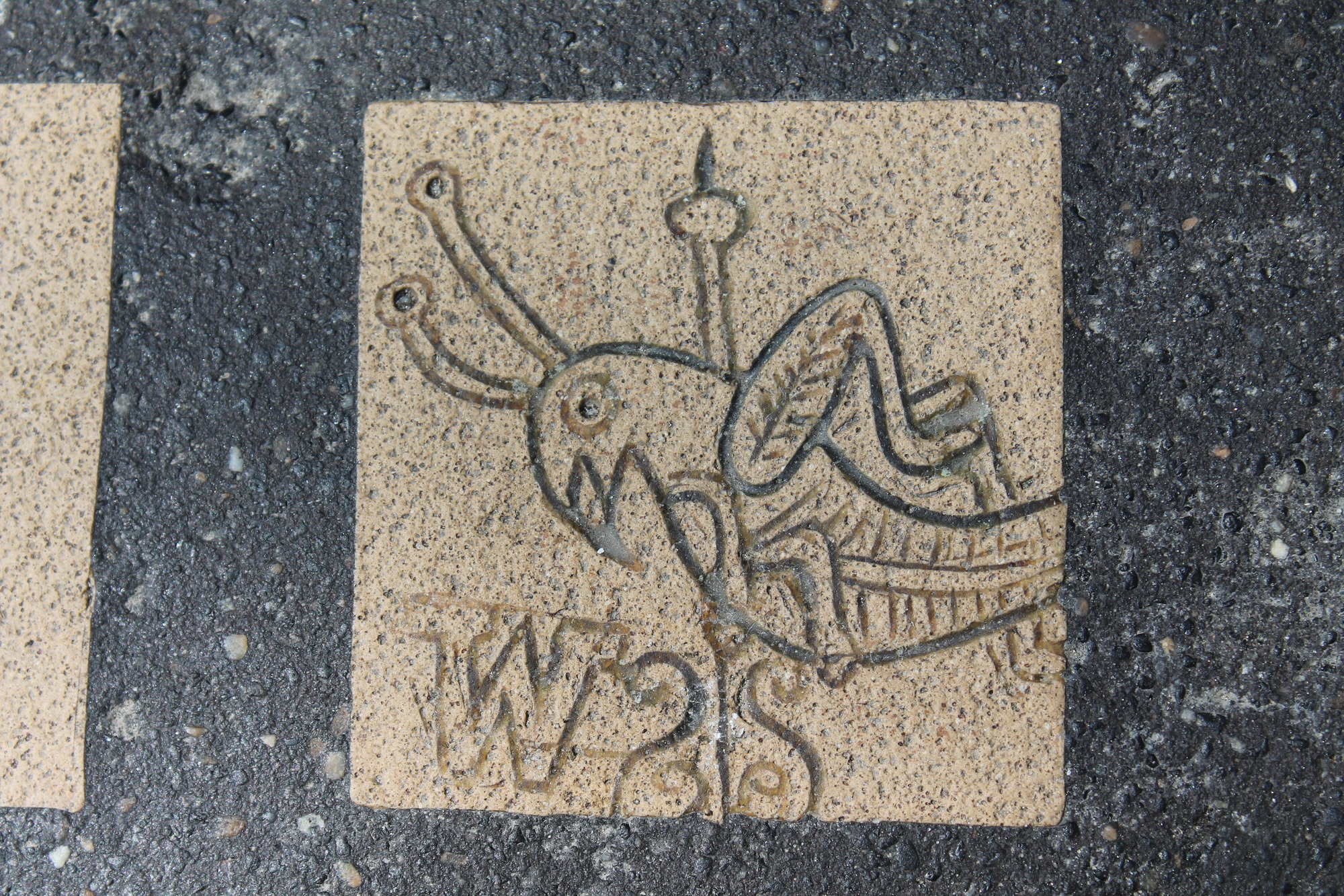 This is a close up of a part of the mosaic for the first public school, the Latin School. It depicts the grasshopper weather vane that is located on top of Faneuil Hall. The grasshopper is facing to the left and has his top legs raised like he is jumping. There is a pole shown going through his body. At the bottom left of the image is a "W" indicating that west is in that direction.