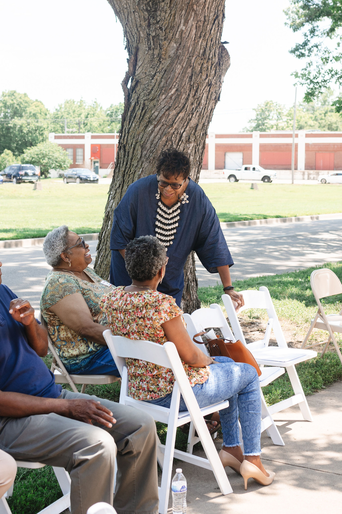 An African American Woman in a blue Dress with a shell necklace, conversates with her two African american friends as she pulls up a seat to join them in watching the Homecoming performances.