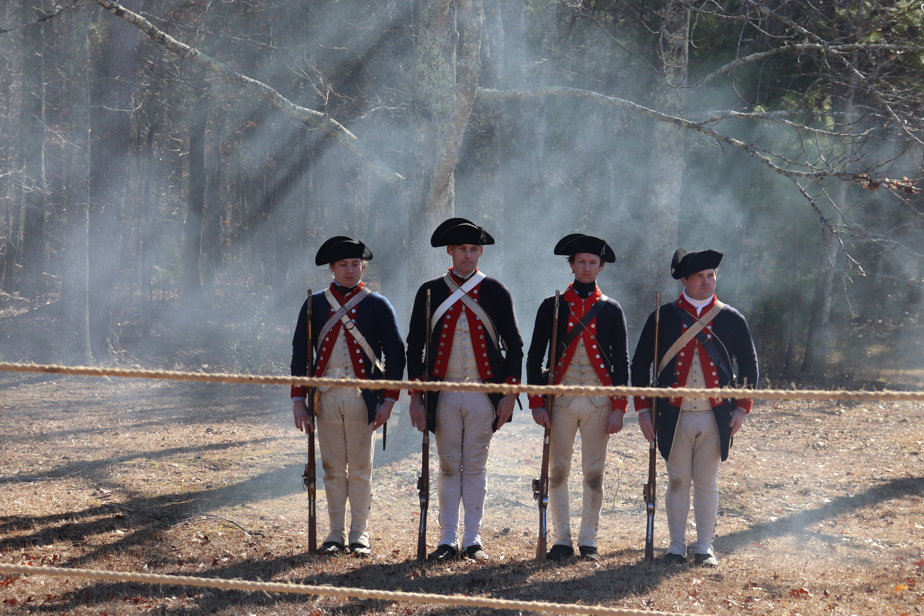 Four men dressed as continental soldiers stand at attention with muskets at their sides in a cloud of gun smoke. 