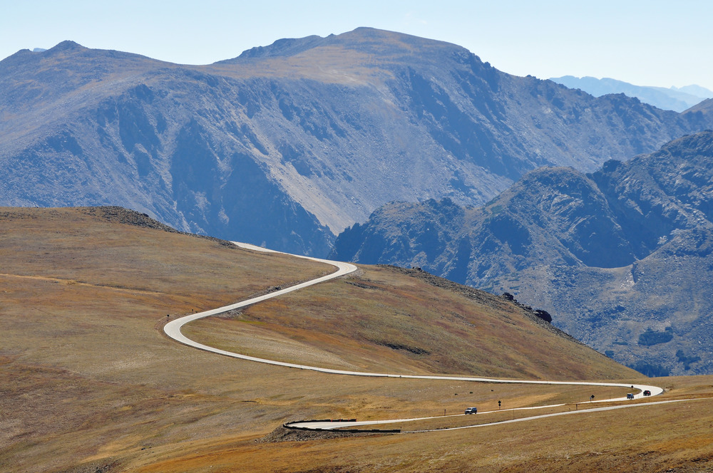 Cars drive a small road dominated by treeless rocky mountains