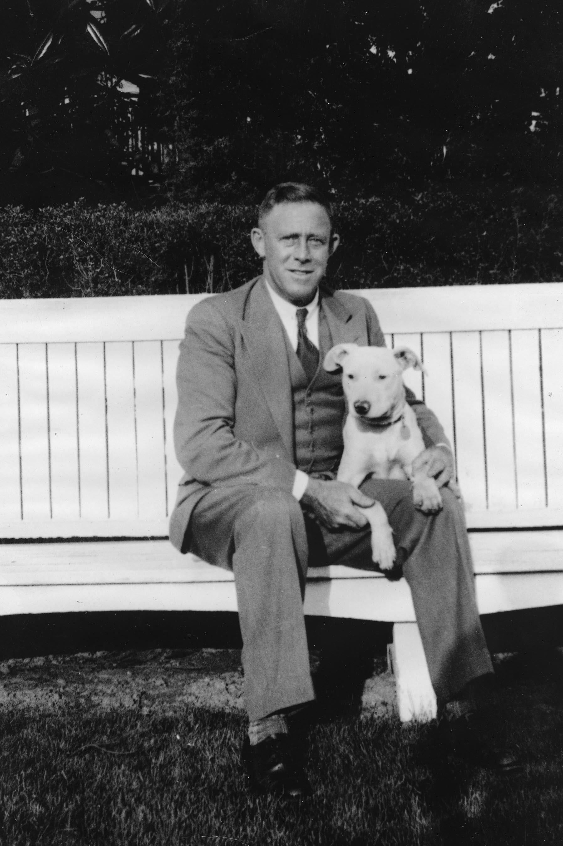 Man wearing a three piece suit and seated on a bench with a dog.