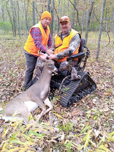 Two men in blaze orange, one in a tracked powered mobility device pose with a buck deer harvested. 