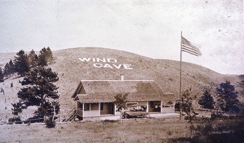 a small building with a car in front of a tall grassy hill, the hill has white lettering that reads 'wind cave'