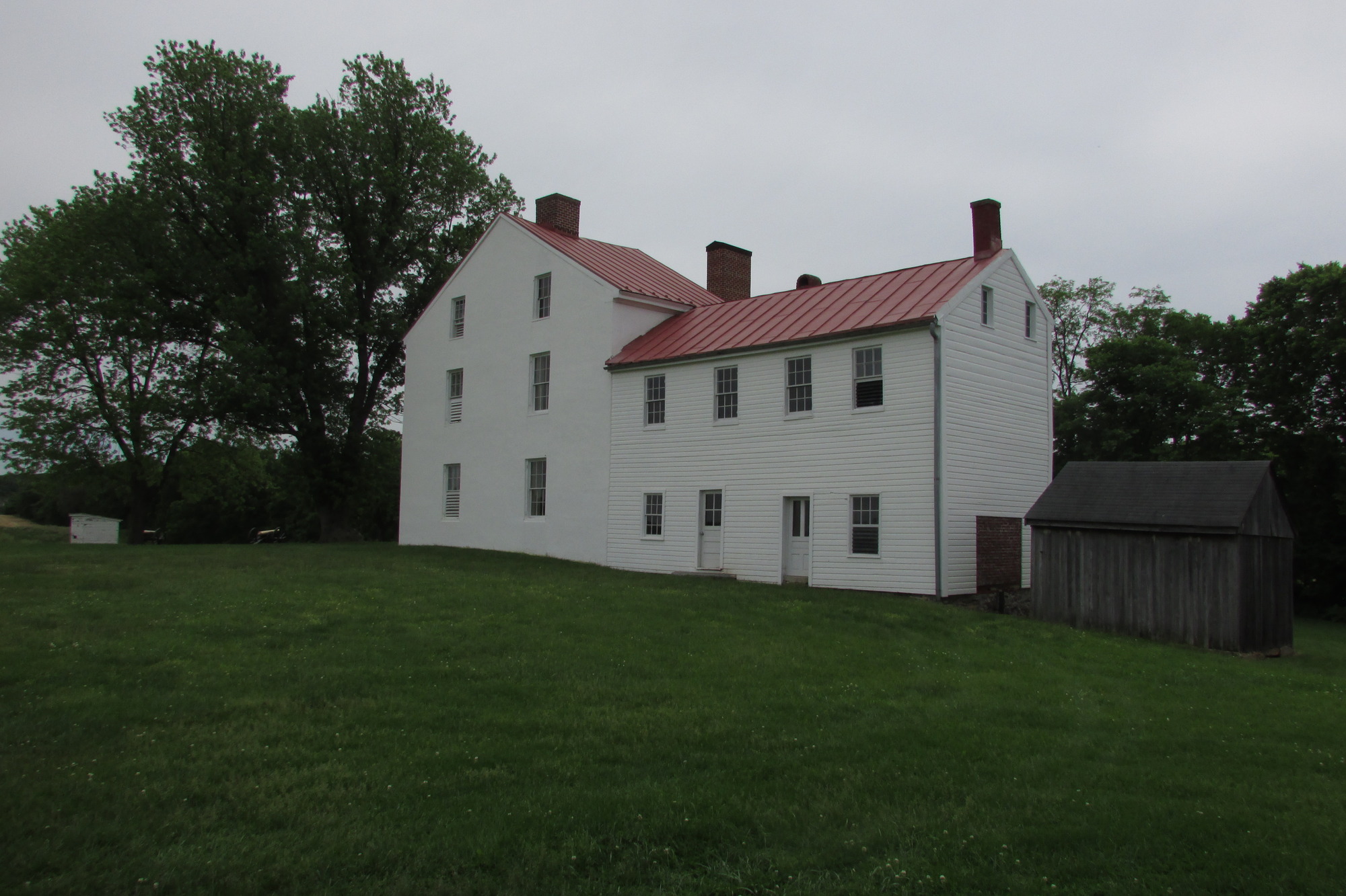 A narrow two-story section of a house abuts a taller section. The roof angles are perpendicular to each other, and rows of rectangular windows line the sides of the structure. It is positioned on a sloping grassy area with trees in the background.