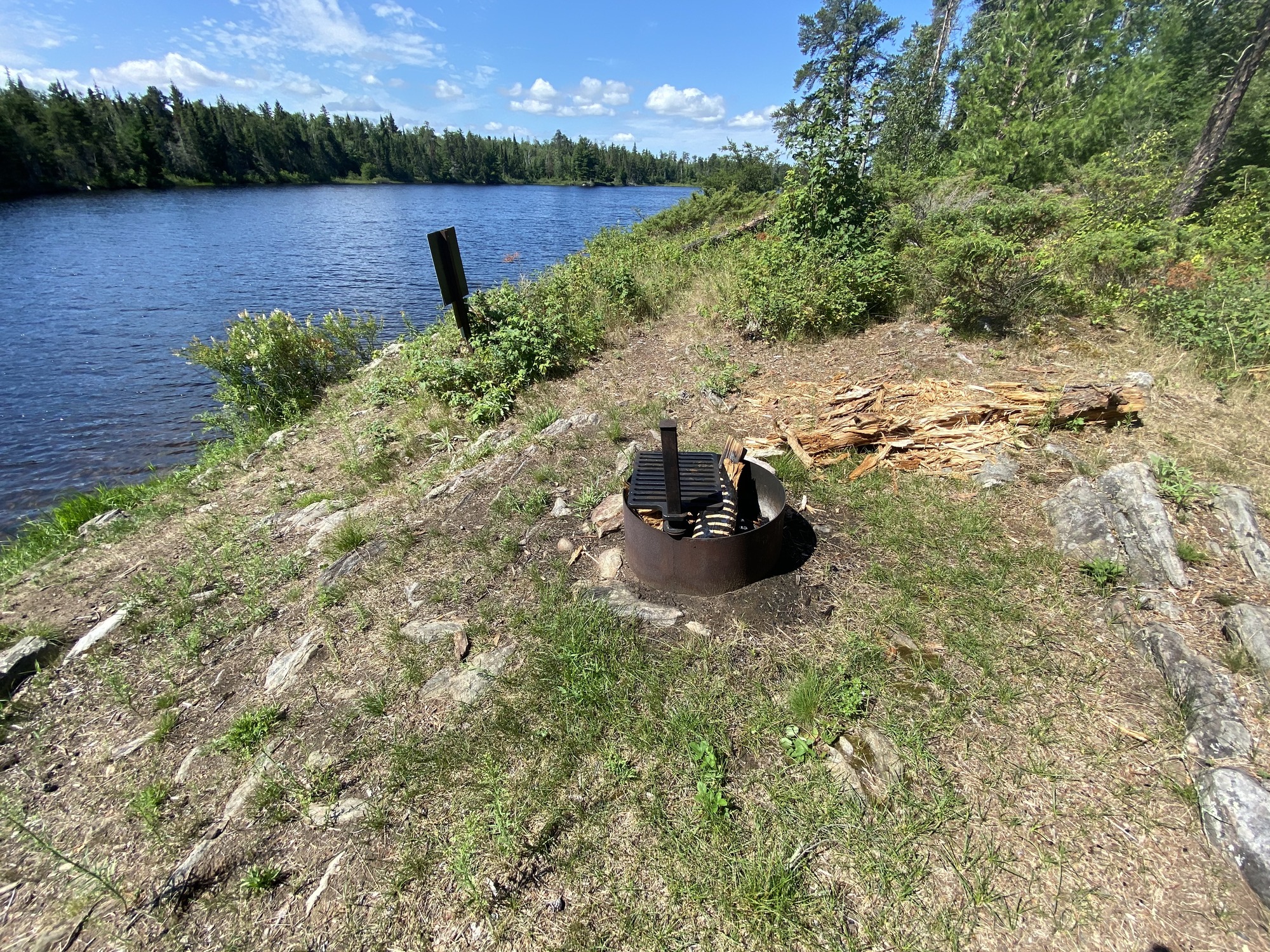 View of the fire ring in the middle of the image with a smash log next to it. The rocky shoreline is down below houseboat site core with a tree shoreline across the water in the background.
