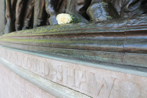The photo shows where the metal of the Robert Gould Shaw Massachusetts 54th Regiment Memorial meets the stone part of the Memorial. Taken at a low angel from the right side, there are multiple feet of men and the horse in metal. In the middle of the image is a white rose laid among the feet. Below that is the stone work, which is a light pink color and has Robert Gould Shaw etched in it.