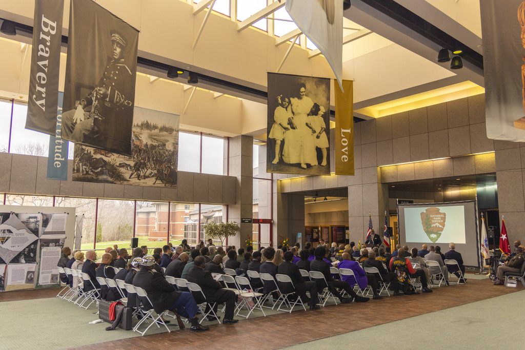 Several people sitting in chairs in a large hall facing toward the right where a man in a dark-colored suit stands at a podium speaking into a microphone.