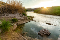 A setting sun illuminates ripples in the Rio Grande and, just above the river, a clear rectangular pool surrounded by a ledged sediment riverbank.