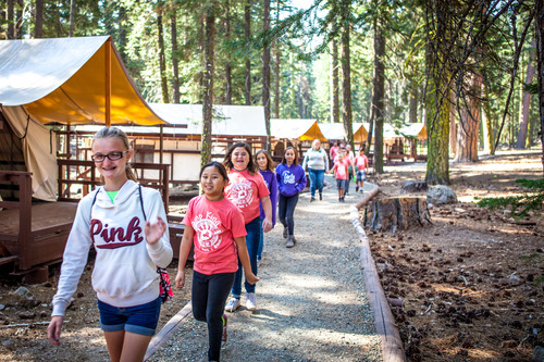 A line of kids walking on a gravel path past canvas camping cabins.