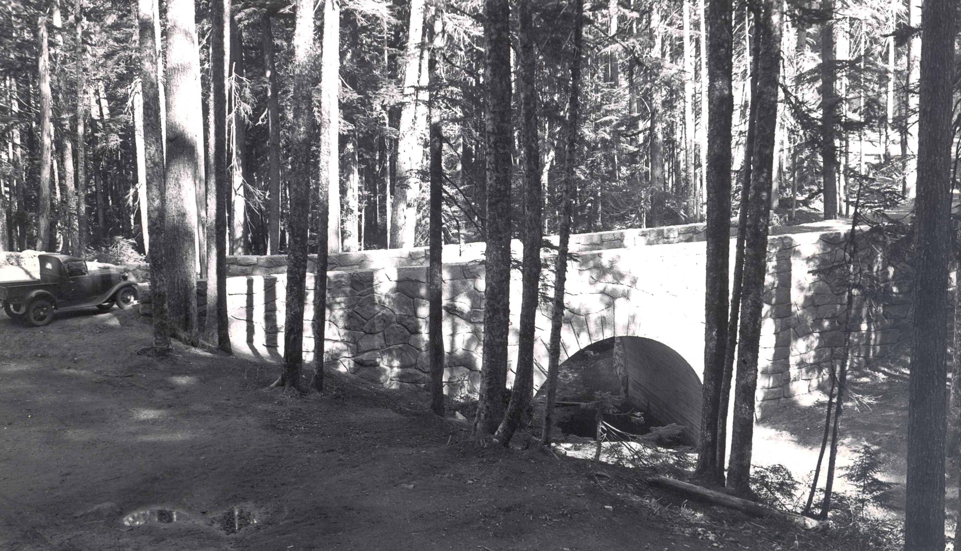 Black and white historic photo of a stone-faced bridge arching over a creek in a forest. An old fashioned car is parked next to the side of the bridge. 