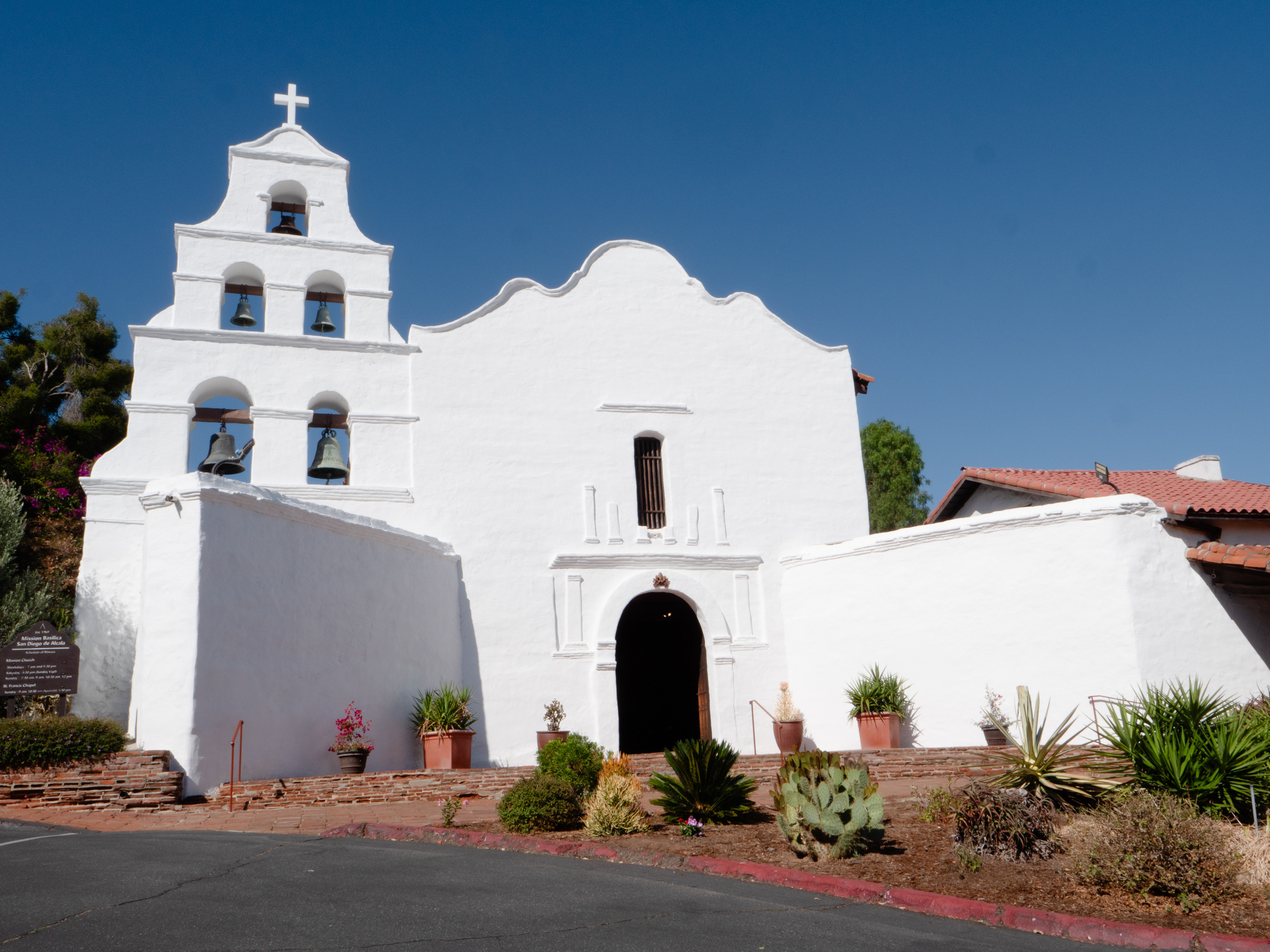 A white facade of a mission with an arched doorway, tower with five bells, and curved roof
