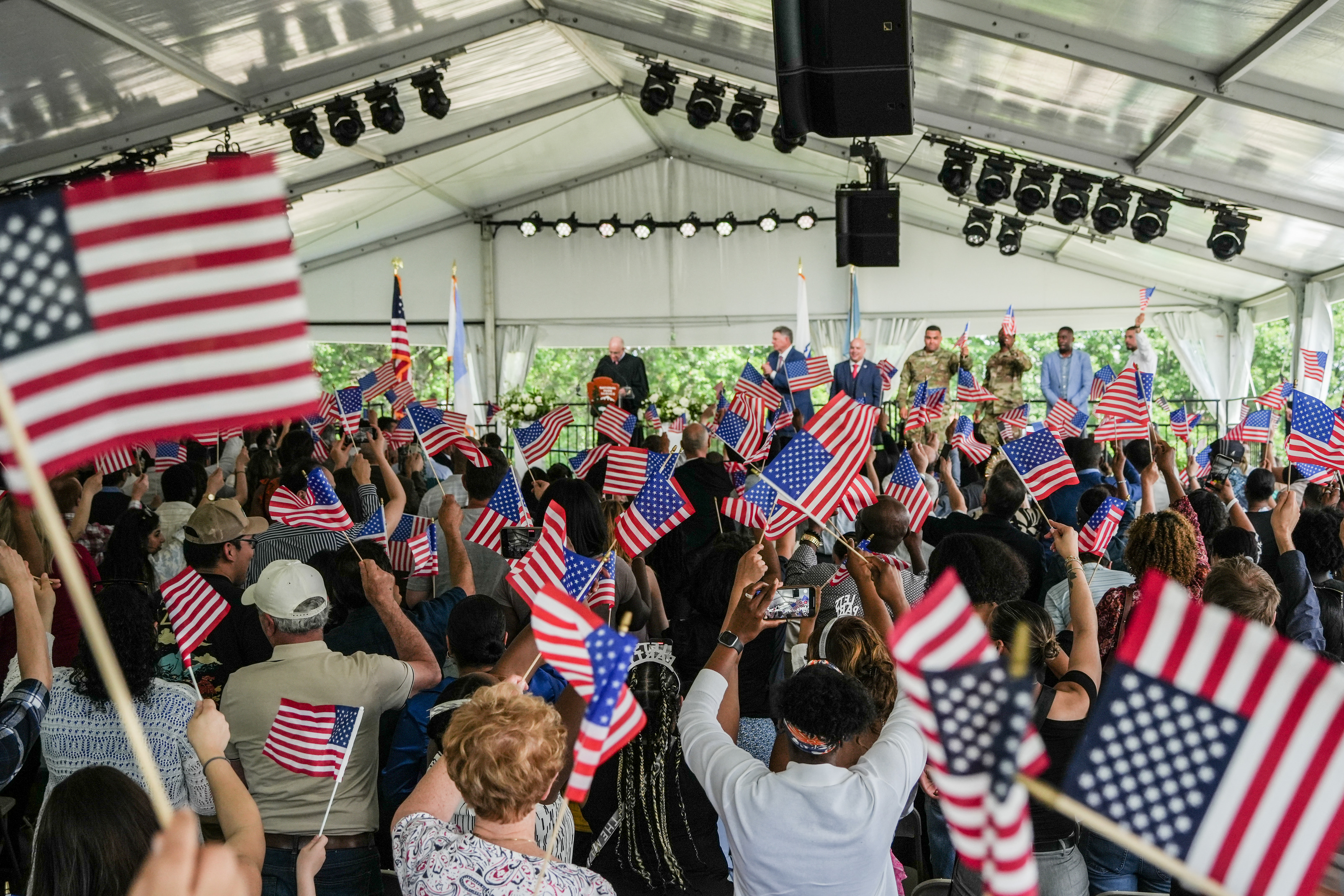 Audience members sitting under a large event tent wave American Flags. 