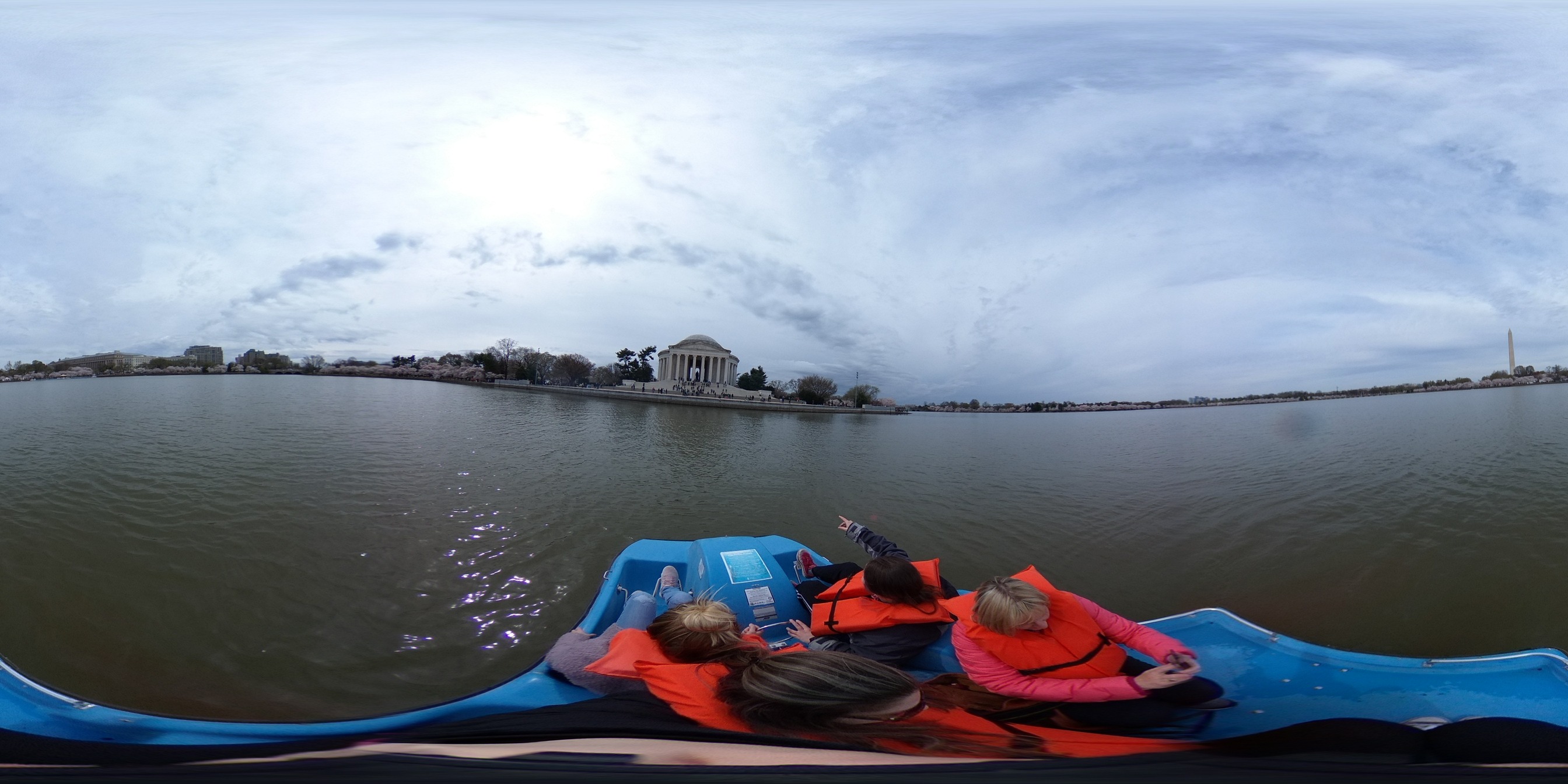Spherical image of four people in a paddle boat in a Tidal Basin surrounded by cherry blossom trees and monuments, including the Thomas Jefferson Memorial, the Martin Luther King, Jr. Memorial, and the Washington Monument.