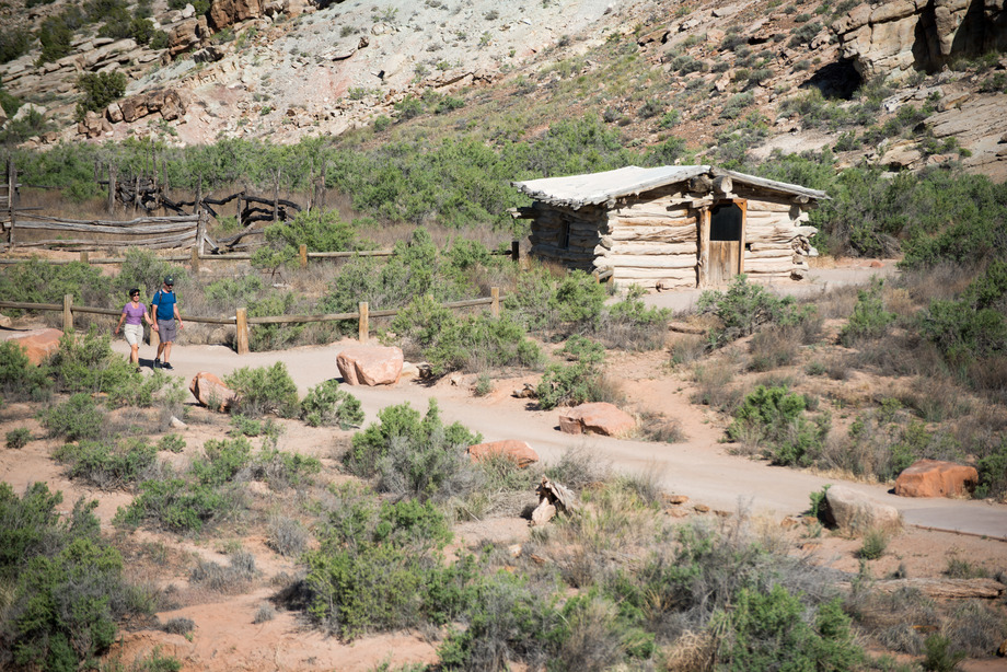 two people walk on a trail passing a log cabin and wood fence