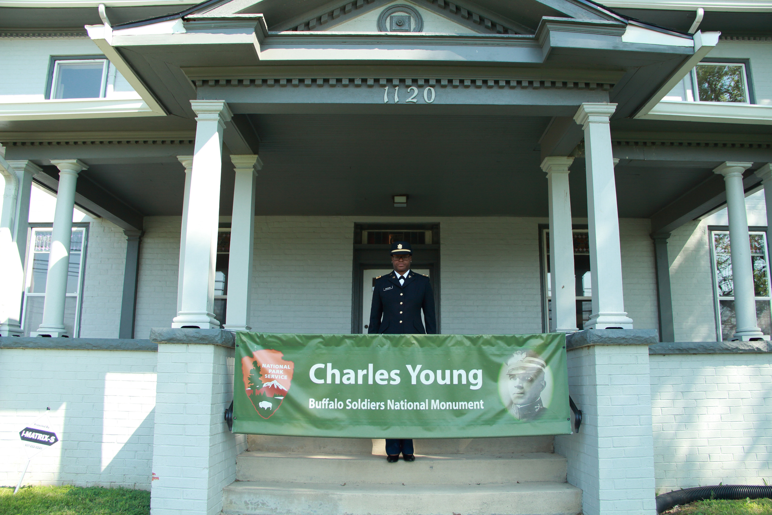 A soldier in a dress uniform stands behind a green banner on concrete steps.