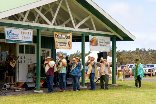 Six men in Hawaiian garments draped over their shoulders blow conch shells and wooden instruments in a traditional Hawaiian ceremony 