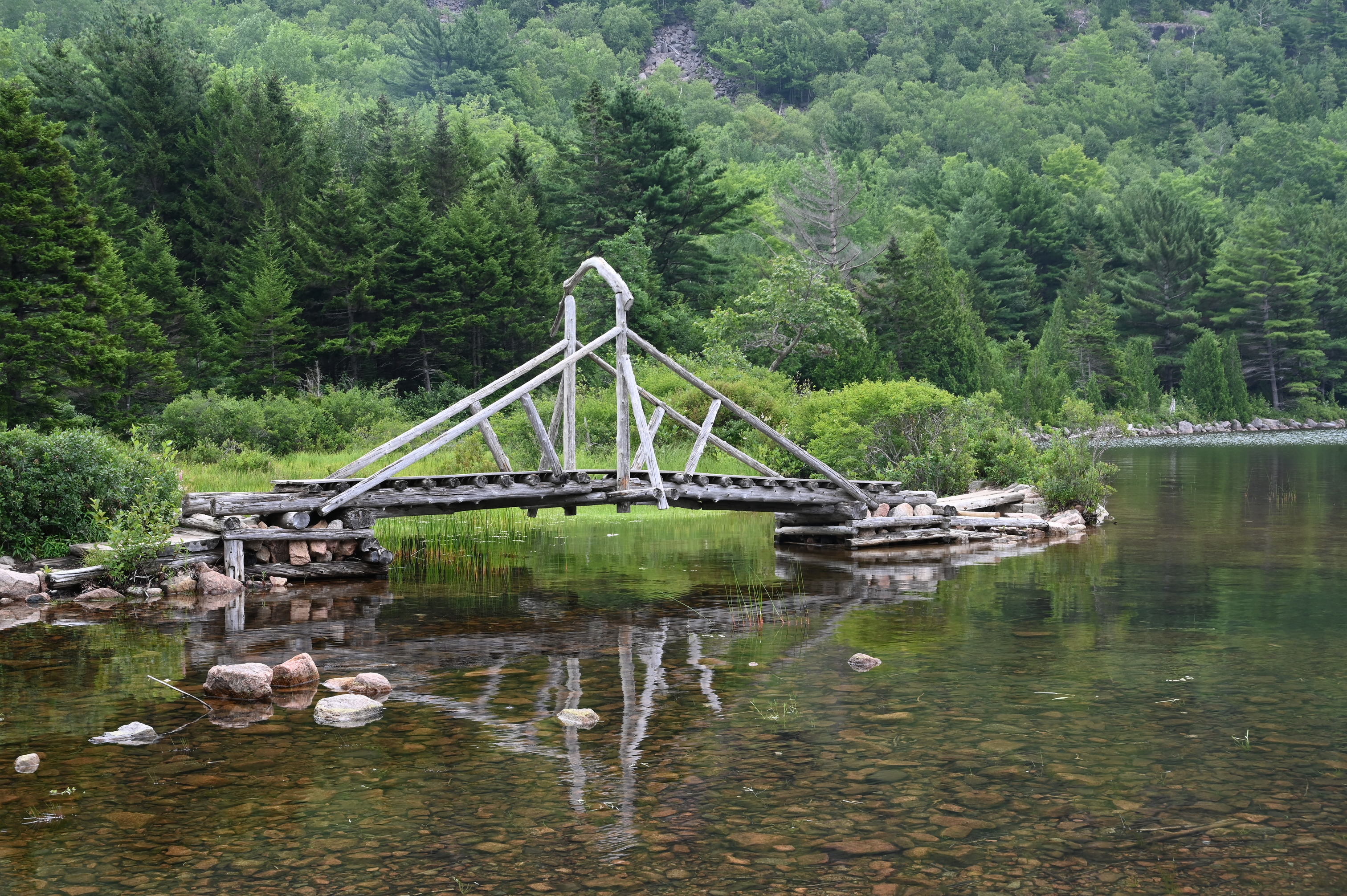A wooden bridge crosses a lake. A mountain and trees are visible behind the bridge.