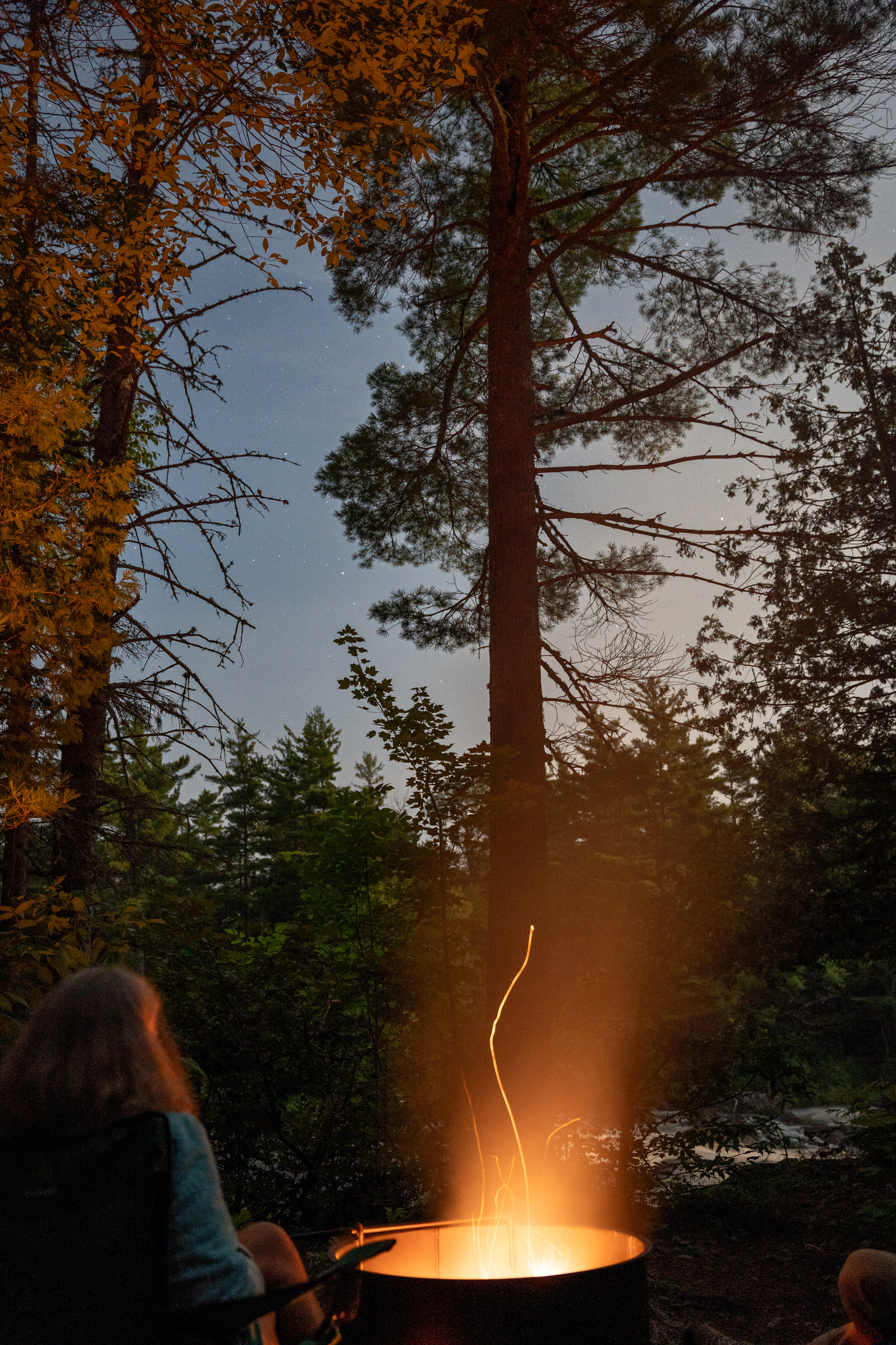 A bright orange fire coming from a fire ring lights up the person sitting in front of it. A river is visible in the background with rapid water flowing while the night sky above showcases stars.