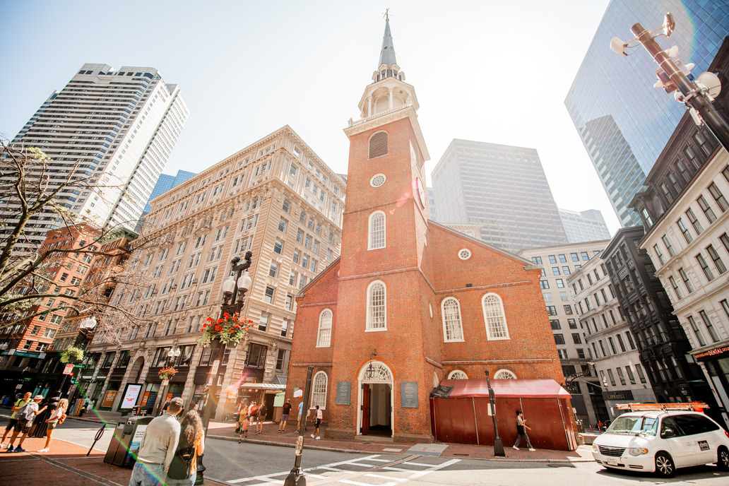 Old South Meeting House, a brick meeting house with a steeple at the front of the building. Sunlight shines brightly behind the Meeting House, providing a slight glare on the camera lens.