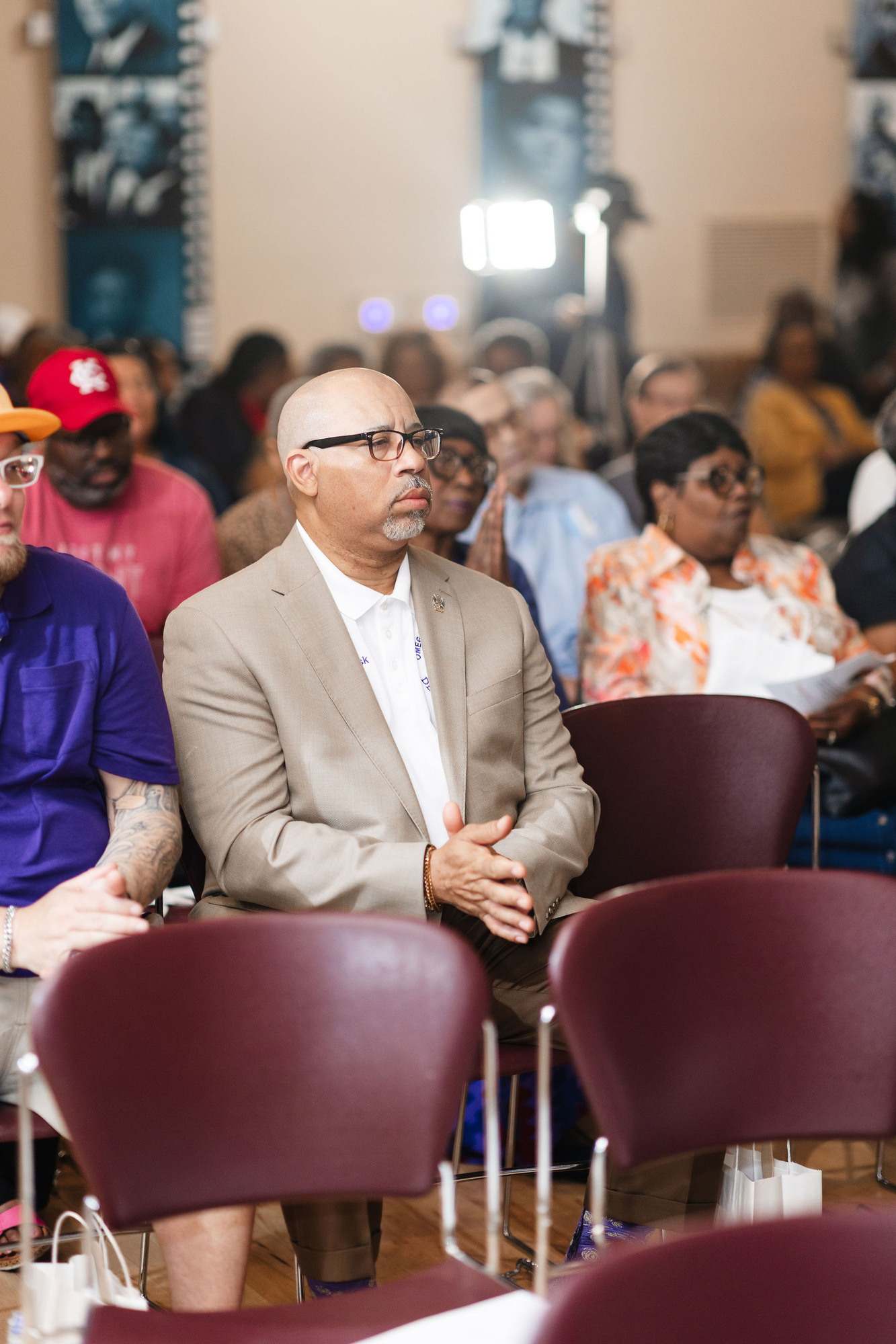 An african american man in a beige suit and black glasses sits on a maroon plastic chair listening intently while surrounded by other african american audience members

