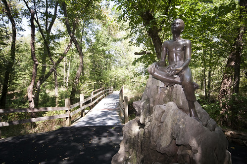 A statue of a young children holding a plant. The statue is one a large stone with the trail bridge in the background. 