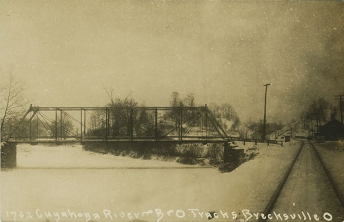 Side view of a metal truss bridge over a frozen river blanketed in snow with railroad tracks and small buildings, right