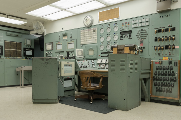 The B Reactor Control Room has faded teal blue paneling covering the walls, which are covered in dials, switches, and buttons. In the center, a wooden rolling chair is surrounded on three sides by additional panels and dials as the central position of the control room. Halogen lights above illuminate the classically nuclear-era-style space.