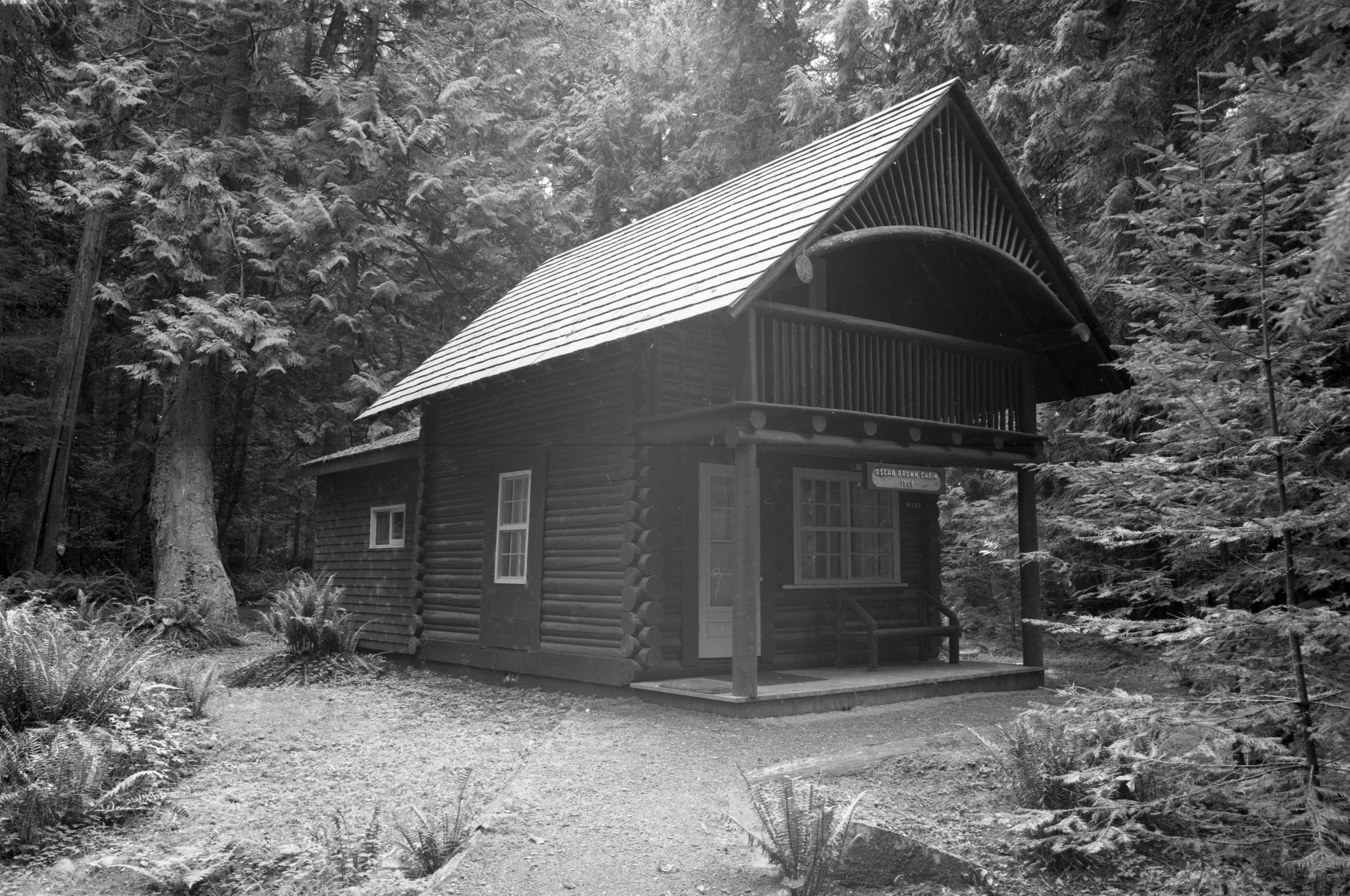 A small wood cabin with a covered porch in a  forest with a dirt path leading up to the porch. 
