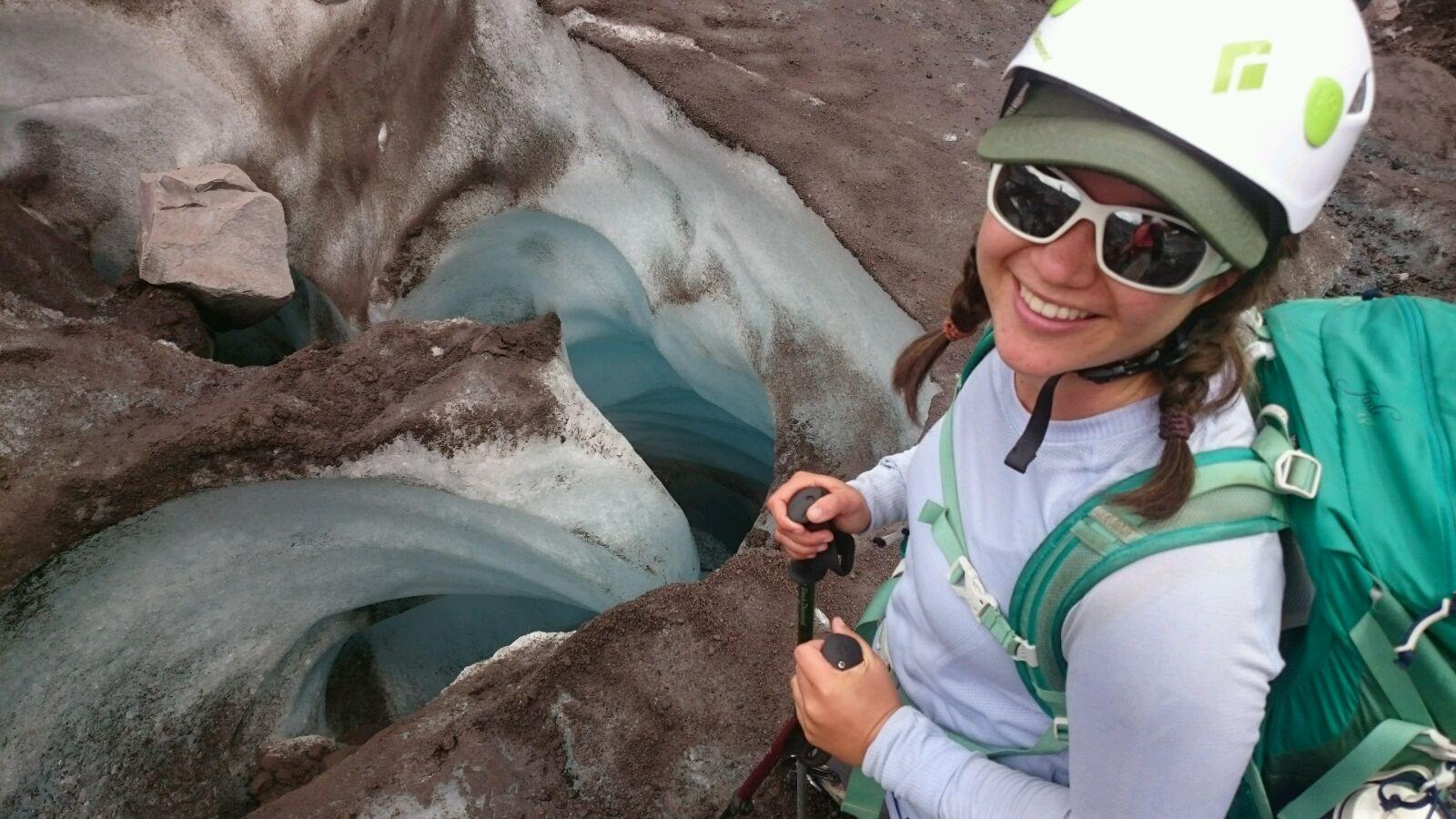a young woman in hiking gear and a white helmet stands on the edge of an ice crevasse.