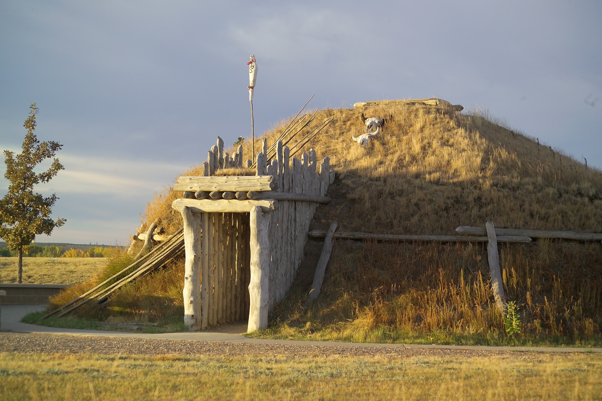 Life-sized earthlodge home with door facing east composed of wooden posts, dirt, and dried grass. 
