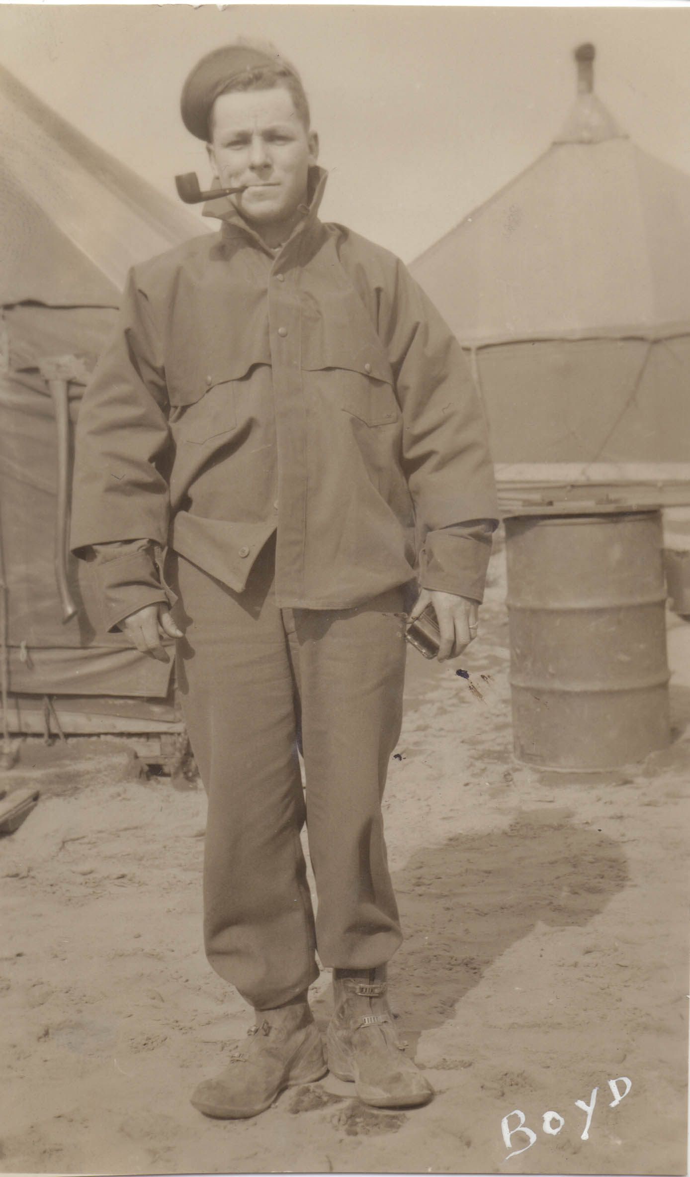 A man with a pipe and jacket collar turned up.  Behind him are canvas tents