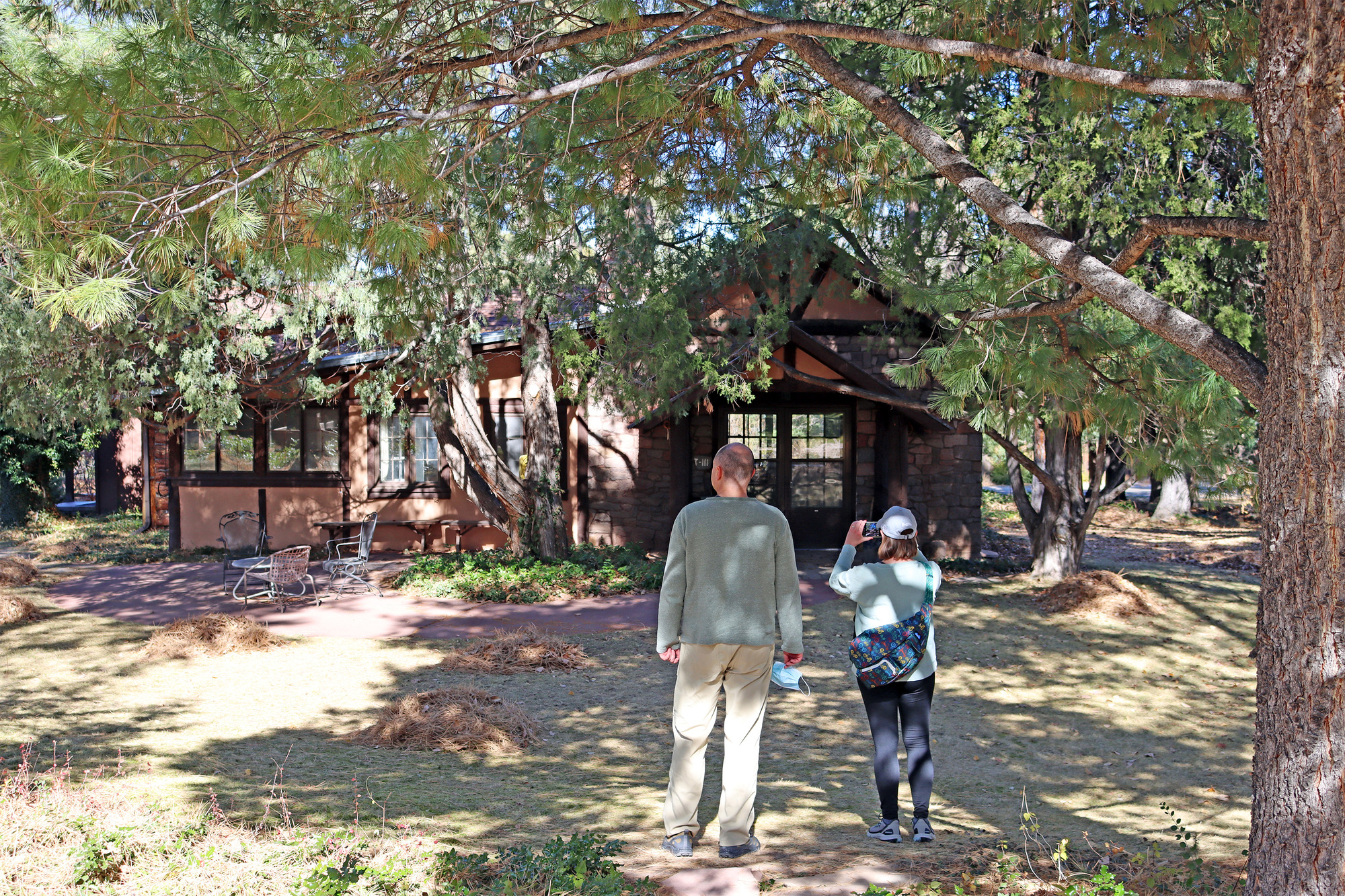 Two people stand in dappled shade under a tree and in front of a single-story historic wooden house that once housed J. Robert Oppenheimer and his family during the Manhattan Project.