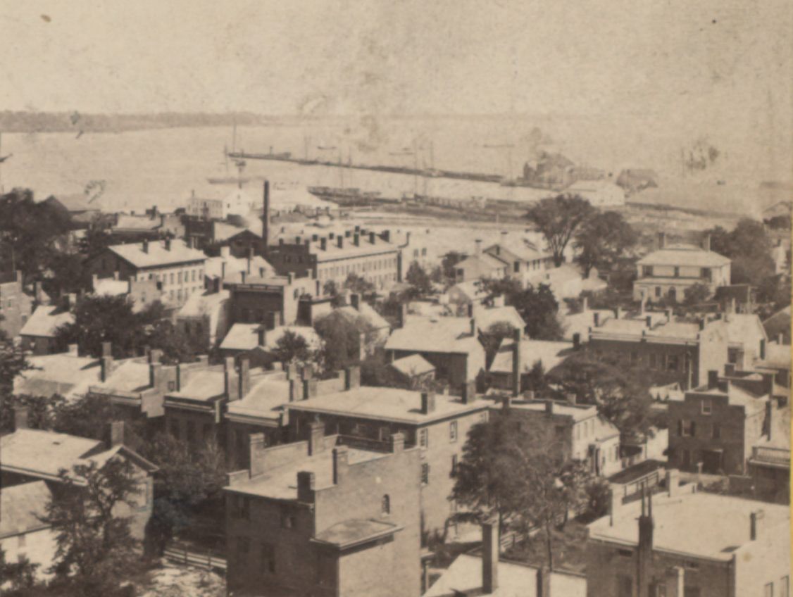 Black and white image overlooking buildings and a harbor in the distance.