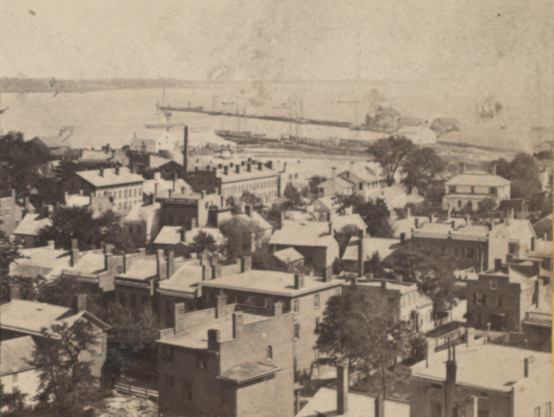 Black and white image overlooking buildings and a harbor in the distance. 