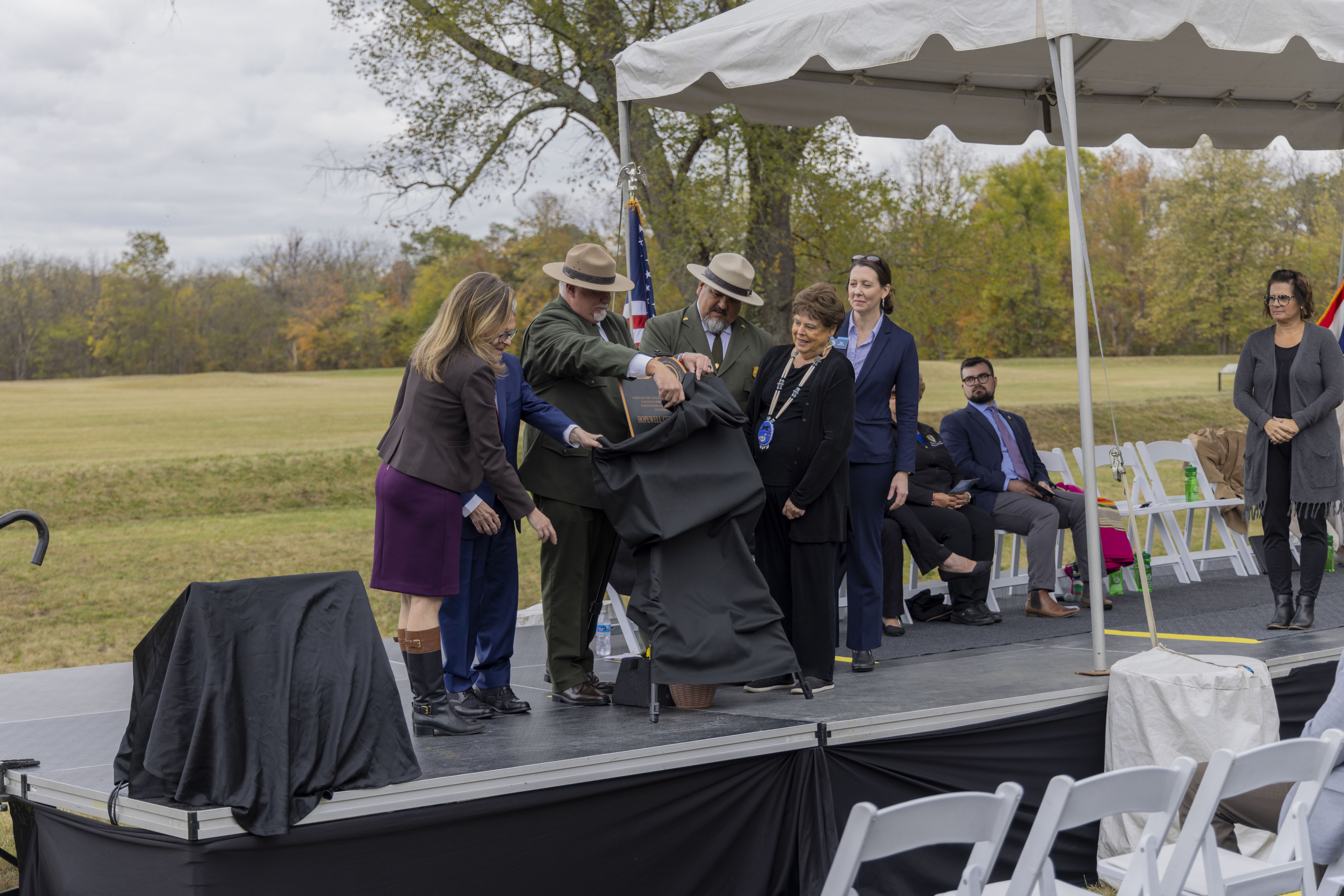 Several people in formal clothing clap while standing behind a black and bronze plaque replica on a stage as they pull off a black cover off of the plaque. Several grass-covered mounds in the background are also seen.
