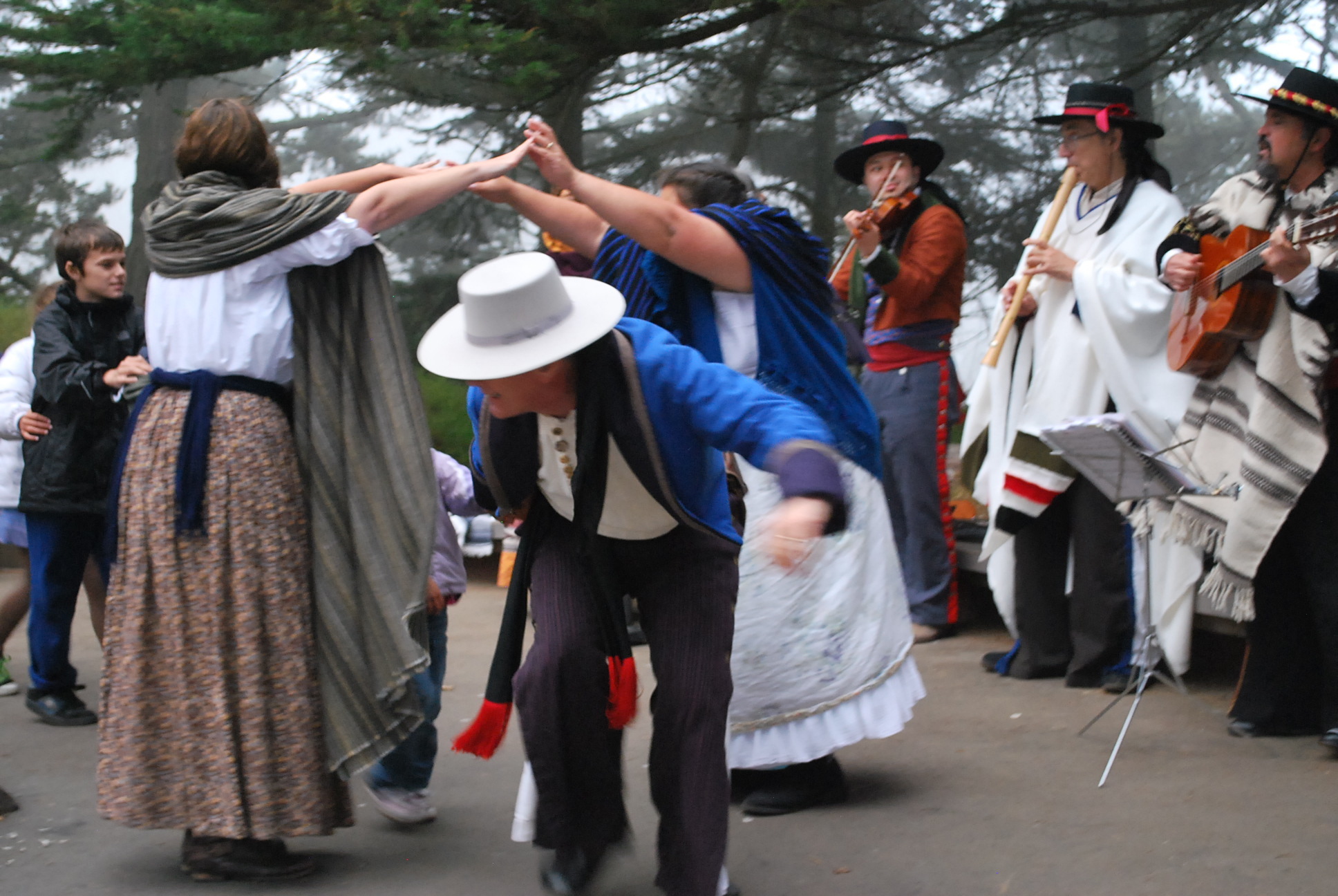 Three people play guitar, violin, and flue while two women join hands to make a tall bridge while people walk under