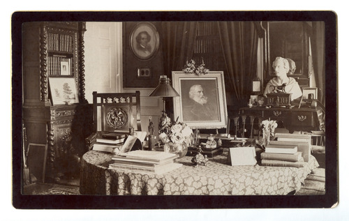 Black and white photograph of top of desk in 19th century study. Shows books, portraits and bust.