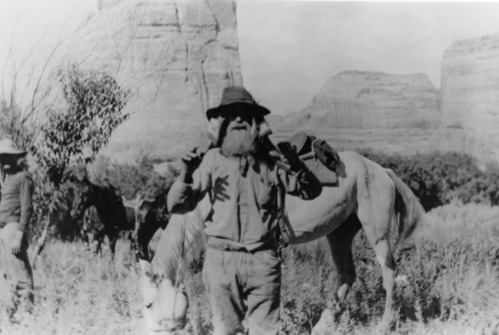 Pat's Hole also known as Echo Park. Pat stands with a horse amongst towering sandstone features. He is protecting his head and neck with a piece of cloth and a floppy hat.