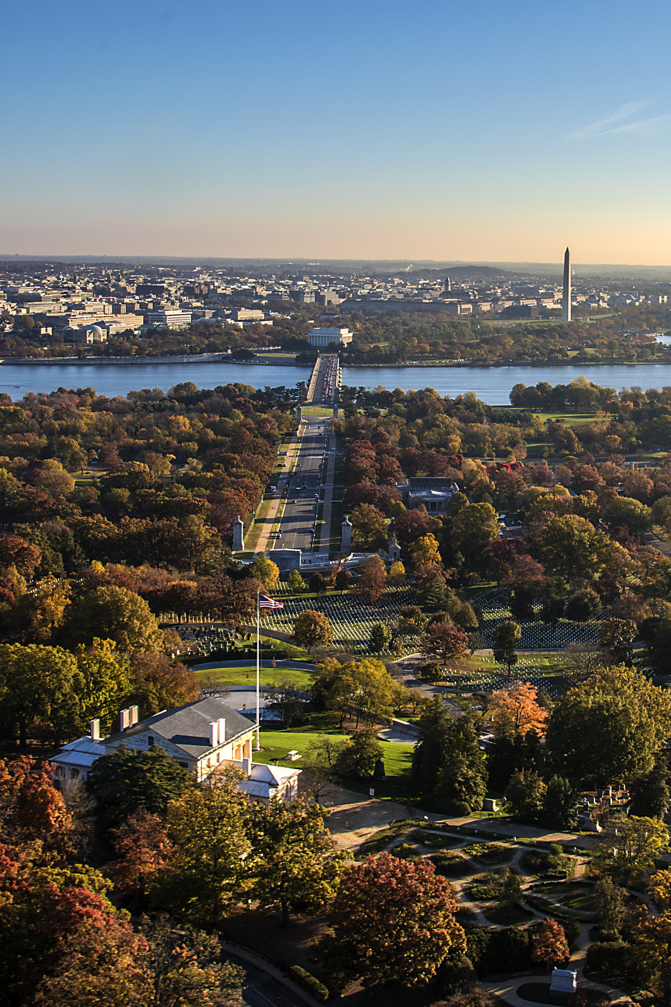 An aerial view with Arlington House and Arlington National Cemetery in the foreground, Arlington Memorial Bridge crossing over the Potomac River, and Lincoln Memorial and Washington Monument visible on the other side of the bridge.