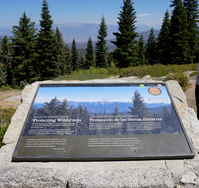 A metal panel sits in a rock base. In the mid ground there are bushes and large trees. In the far background there are large mountains. 