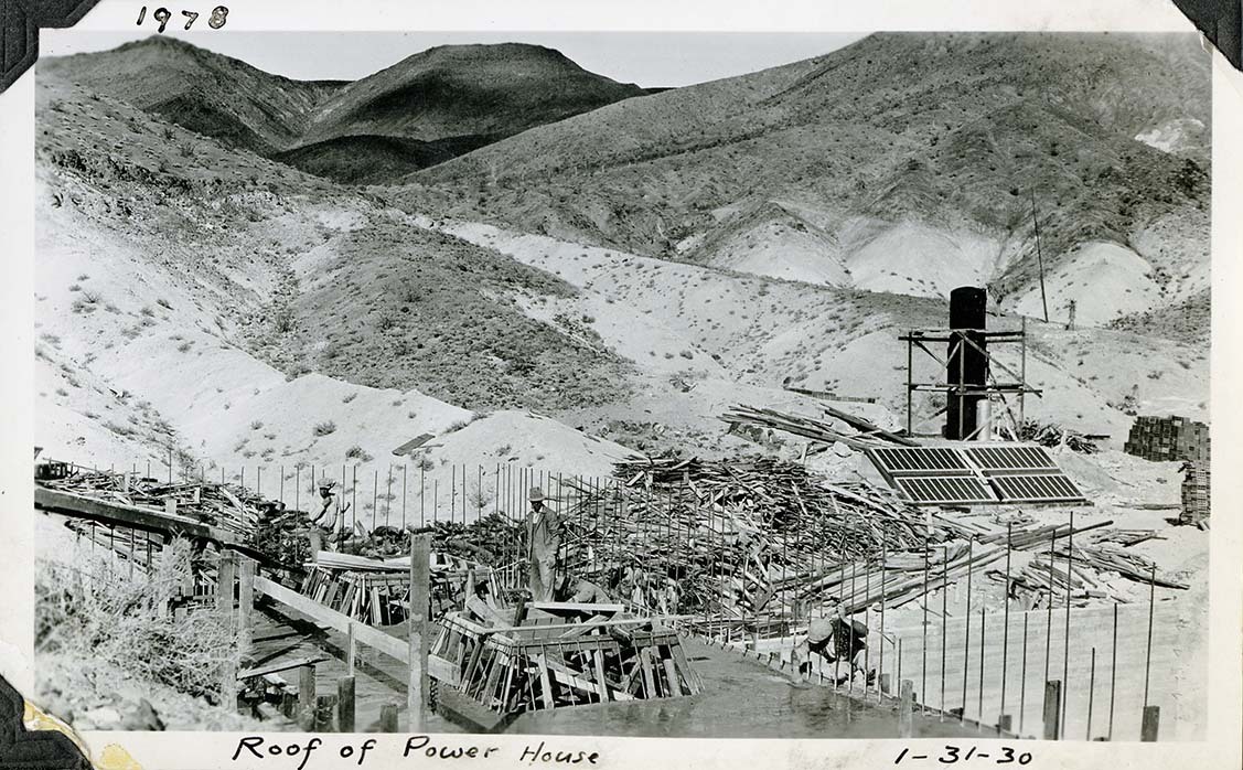 This is an historic black and white photograph from the Scotty's Castle Historic Photograph Collection, Death Valley National Park of four men on newly poured concrete roof. Skylights formed with lumber on roof. Line of rebar extend up from roofline. Huge pile of discarded form-lumber behind. Four solar panels and tall black hot water tank to left. Background filled with slopes of barren desert hillsides. Inscriptions in black ink along upper and lower border.