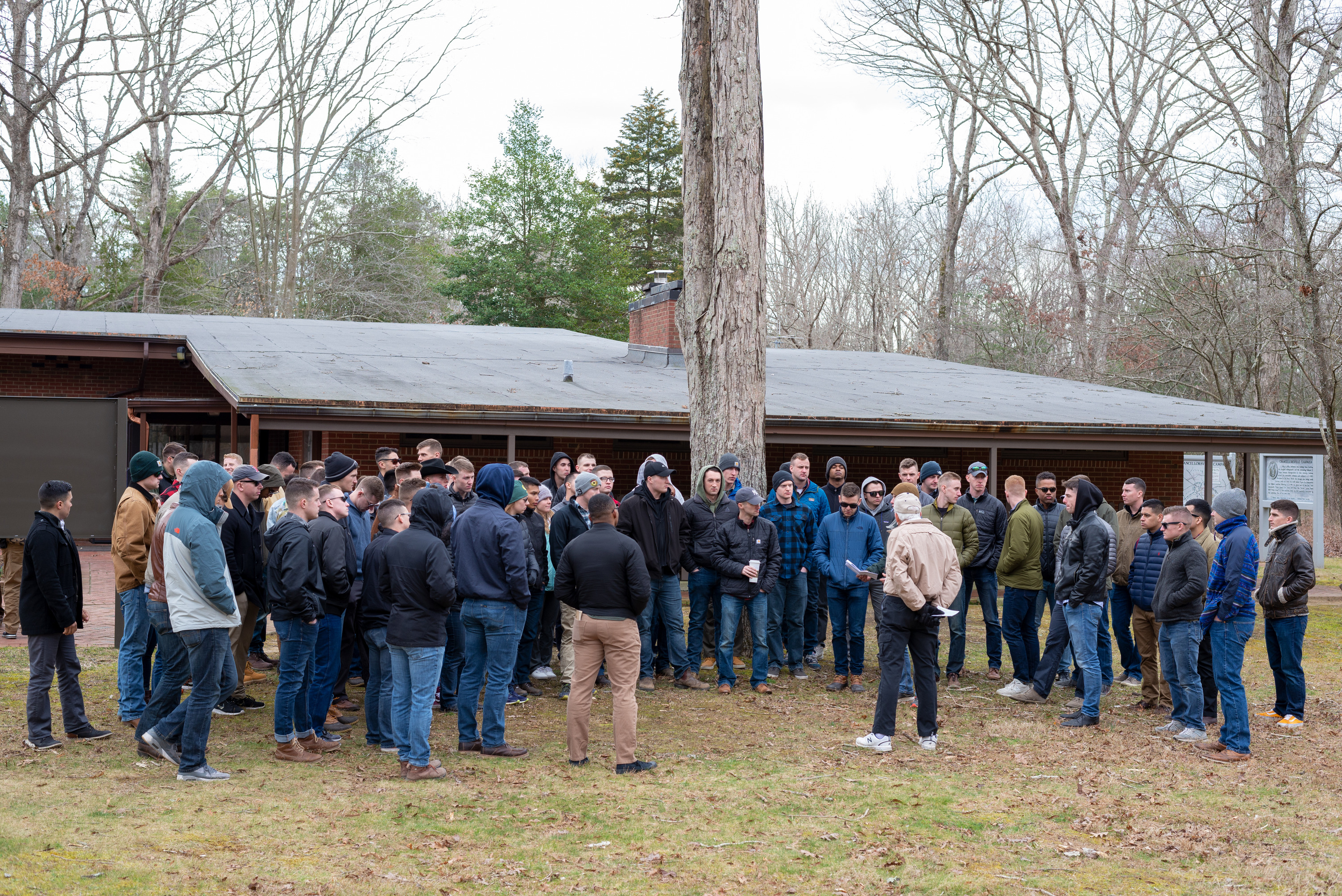 A volunteer gives a talk to a group of people on a path with a brown, single story building in the background.