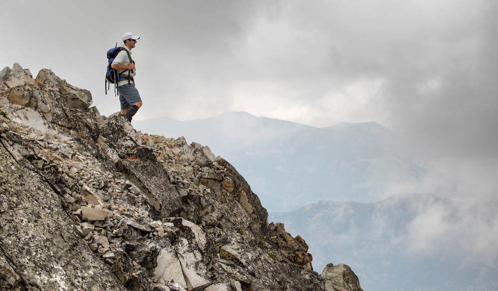 Hiker standing on rocky slope with mountains peaking through clouds in background