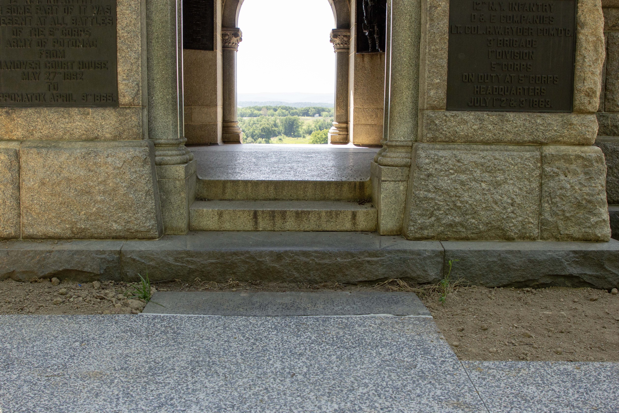 A concrete walkway surrounded by topsoil leads to the stone steps and a doorway arch of a large stone monument, trees can be seen through the other arch of the monument. 