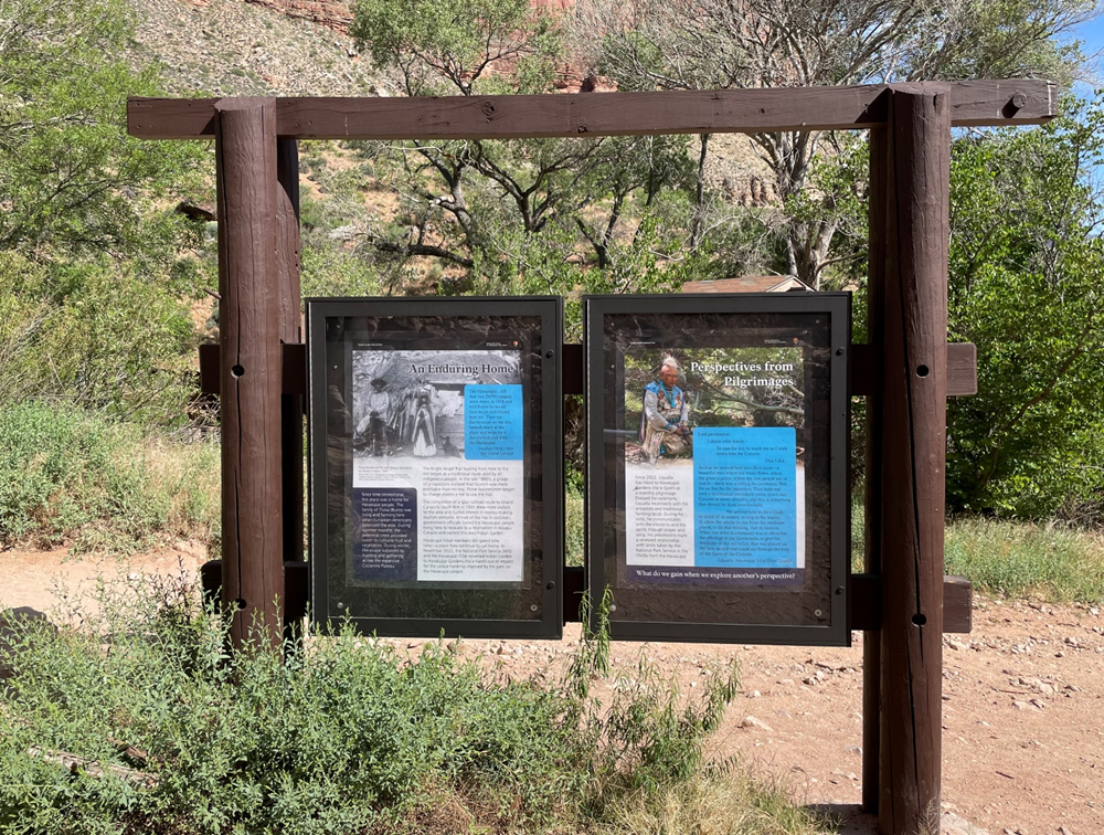 A wooden outdoor signboard displays two informational posters along a dirt trail surrounded by green shrubs and trees, with rocky terrain in the background.
