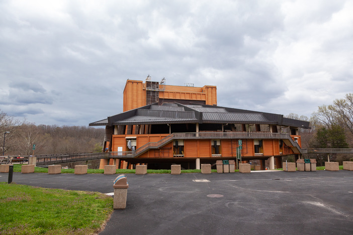 A building with new, bright orange siding is in the center of the frame. White clouds loom overhead. There is a paved sidewalk in front of it and green grass.