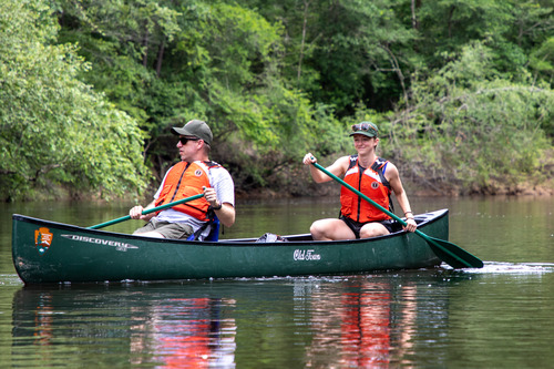 A man, seated in front, and woman, in the rear, paddling a green canoe on a calm river with dense forest on the far bank.